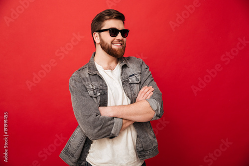Image of fashion young man wearing sunglasses and denim smiling and posing with hands folded, isolated over red background