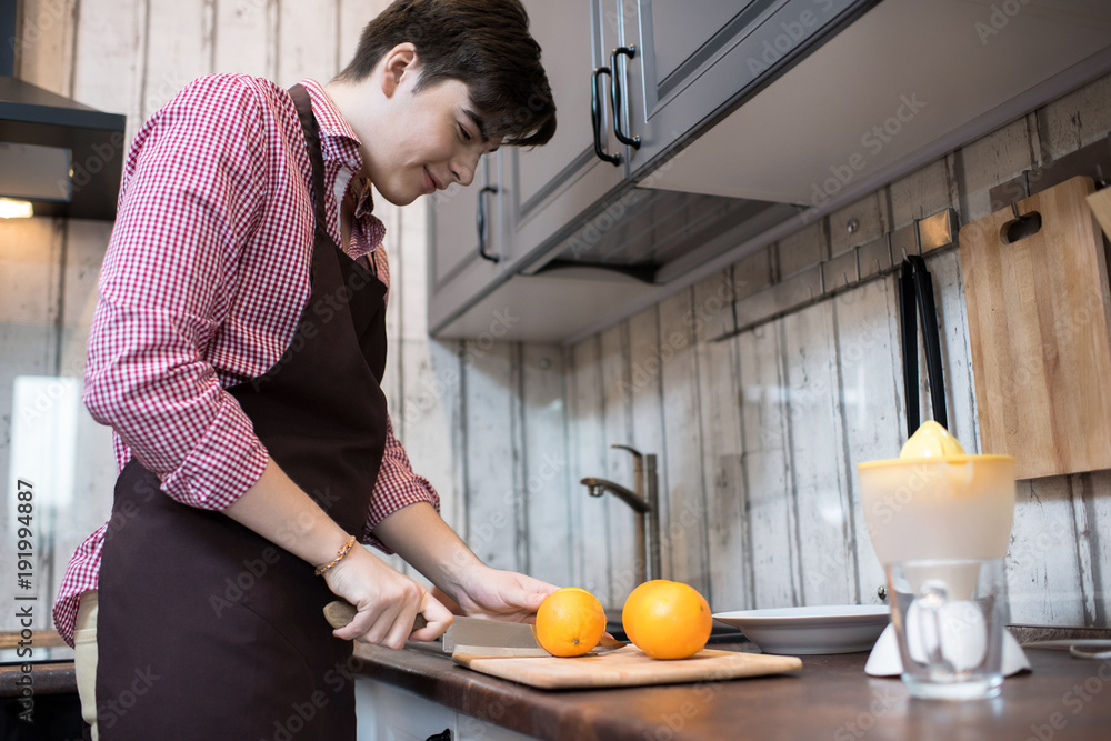Side view portrait of smiling teenage boy cooking alone in modern kitchen, making orange juice, copy space