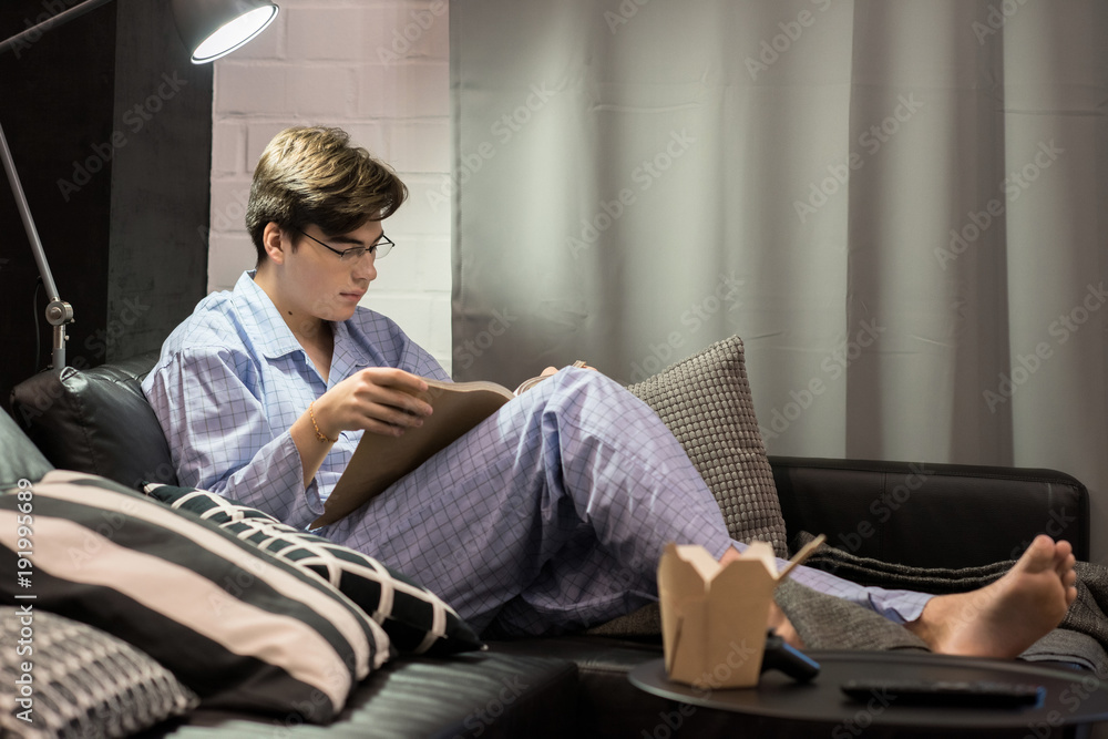 Portrait of teenage boy wearing glasses reading book while chilling at home sitting on bed in pajamas