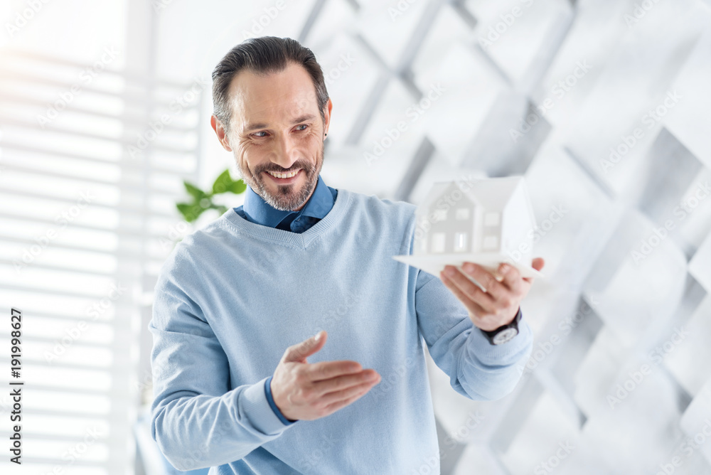 Smiling. Handsome content dark-haired bearded man smiling and holding a model of a future house and looking at it Smiling. Handsome content dark-haired bearded man smiling and holding a model of a future house and looking at it