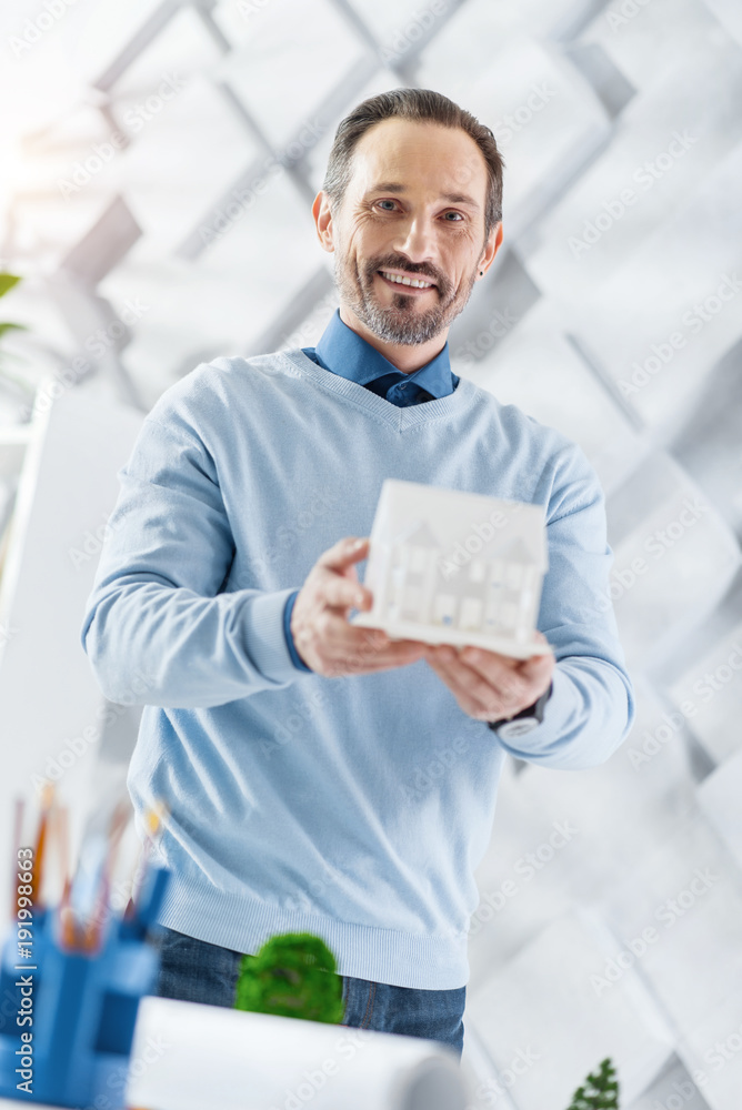 Good job. Good-looking alert dark-haired bearded man smiling and holding a model of a future house while standing at the table