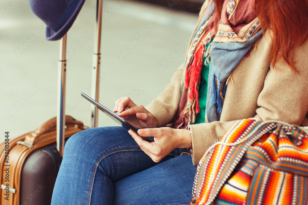 Hipster tourist texting message on tablet or technology mockup. Person traveler using computer on train station background close. Female hands holding gadget on blurred backdrop. Luggage