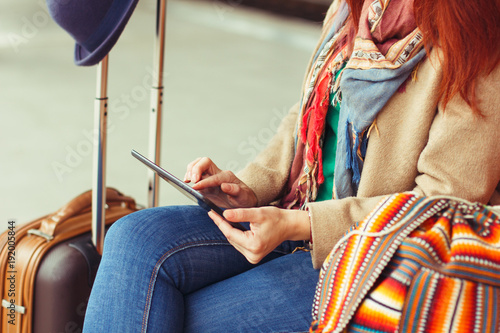 Hipster tourist texting message on tablet or technology mockup. Person traveler using computer on train station background close. Female hands holding gadget on blurred backdrop. Luggage