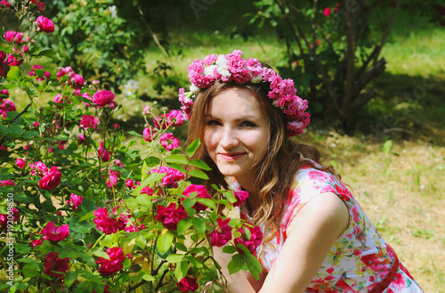 Beautiful girl in pink flower crown sits near the rose bush in green summer park. Pink floral dress. Spring woman photo. International women's day and mother's day holidays. Flower chaplet.