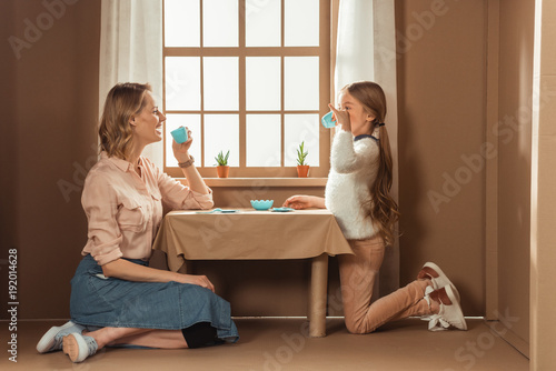 laughing mother and daughter having tea party in cardboard house