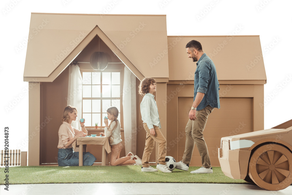 Φωτογραφία young father teaching his som to play soccer on yard of cardboard house isolated