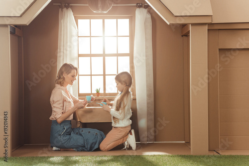 mother and daughter having tea party in cardboard house against window