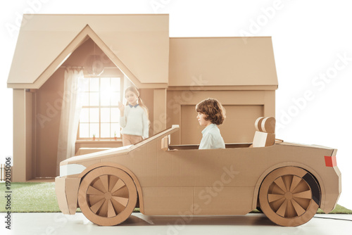 kid riding cardboard car in front of house while girlfriend waving to him isolated on white photo