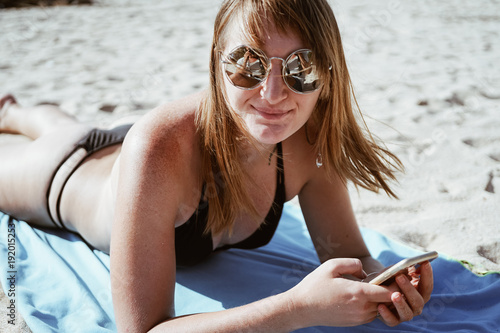 Portrait of a redhead girl in round mirror sunglasses. The girl lies on the sand near the ocean.