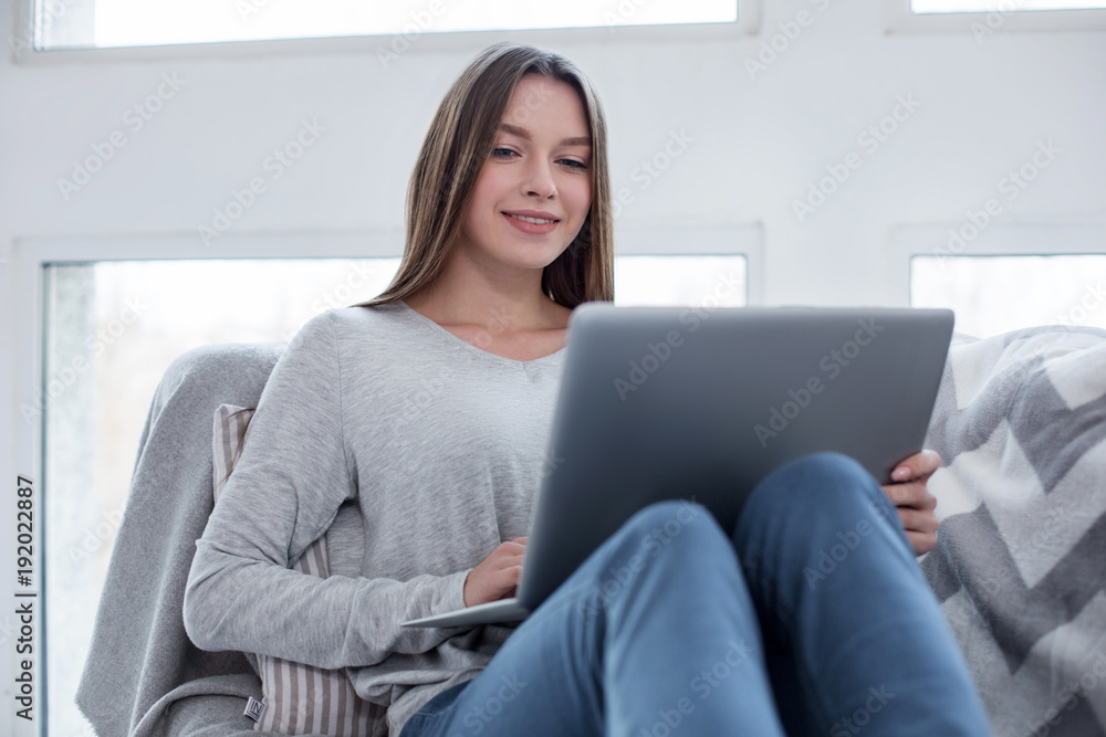 Naklejka premium Weekend. Good-looking exuberant long-haired slim girl smiling and sitting on the sofa and working on her laptop