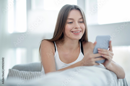 Chatting with friends. Pretty joyful dark-haired young woman smiling and sitting on the sofa and typing on her phone