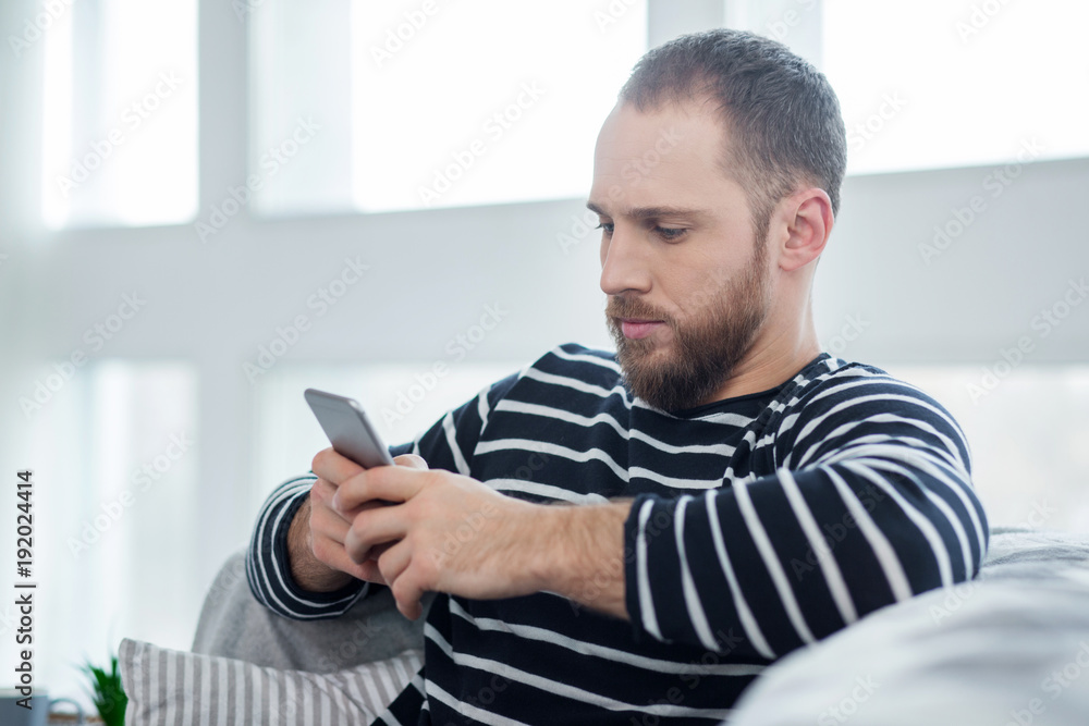 Seriousness. Handsome concentrated bearded young man smiling and sitting on the couch and typing on his phone