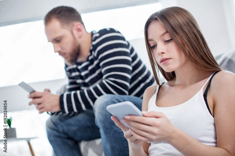 Concentration. Beautiful concentrated long-haired woman holding her phone and her man sitting on the couch and using his phone Concentration. Beautiful concentrated long-haired woman holding her phone and her man sitting on the couch and using his phone