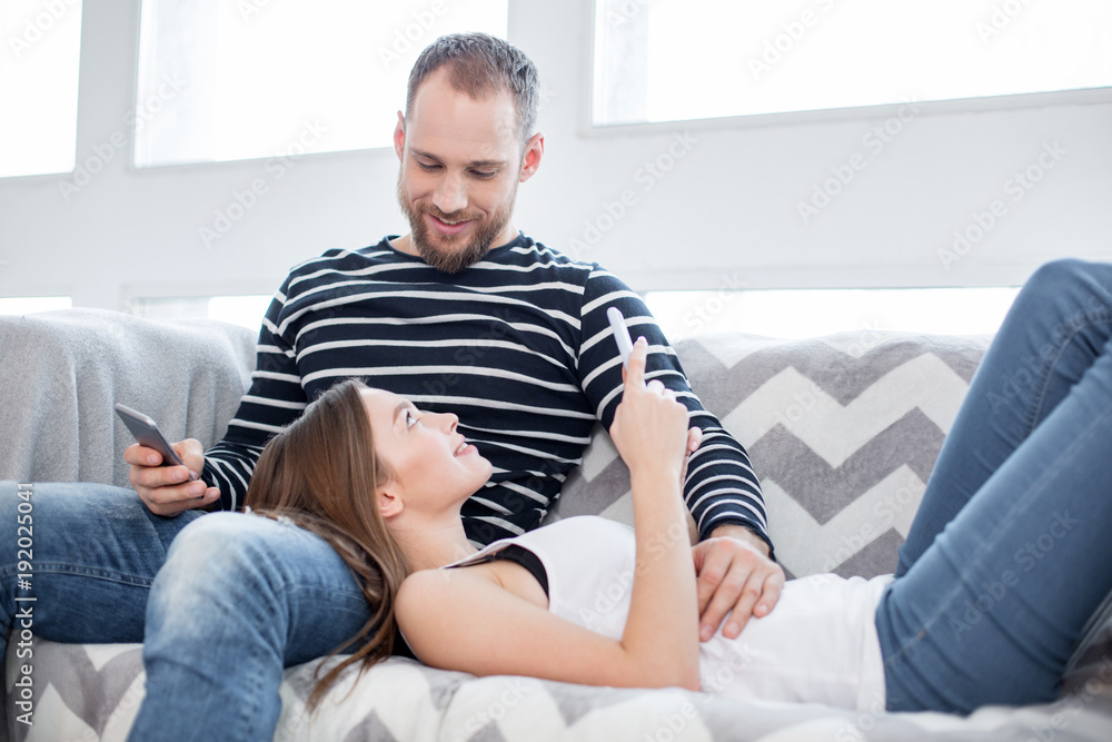 My darling. Good-looking content bearded young man smiling and sitting on the couch with his phone and his girlfriend lying on his lap with her phone
