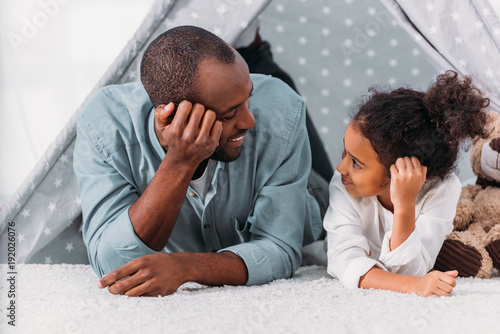 african american father and daughter lying on floor and looking at each other at home