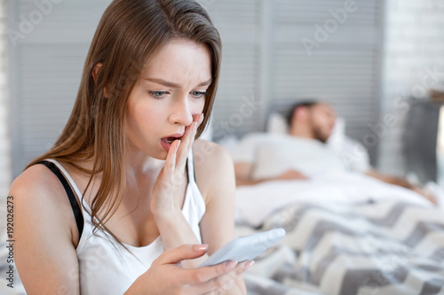 Oh my god. Pretty startled heart-broken woman reading her mans messages while he sleeping and she sitting on the bed photo