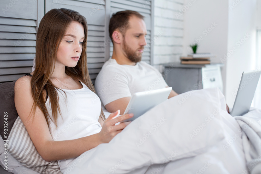 Our family. Attractive concentrated long-haired young woman using her tablet and her man using his laptop while lying in bed Our family. Attractive concentrated long-haired young woman using her tablet and her man using his laptop while lying in bed