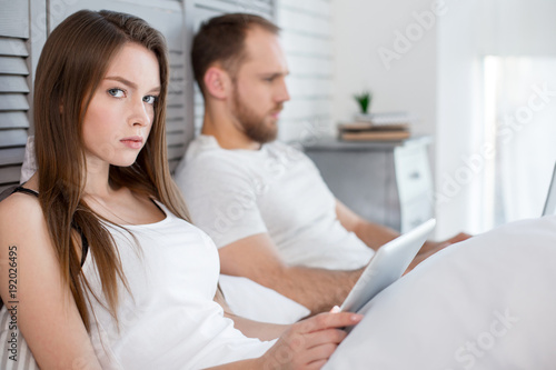 Dissatisfaction. Attractive angry long-haired young woman using her tablet and her man lying in bed next to her and paying no attention to her