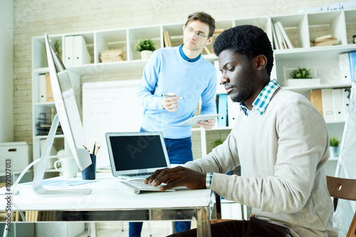 Portrait of two young specialists, one of them African-American, working together in modern office discussing projects while sitting at desk and using PC