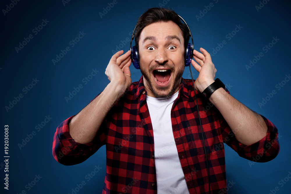 Handsome bearded man in red shirt isolated on dark blue background listening to music in his headphones on his phone and singing along with joy