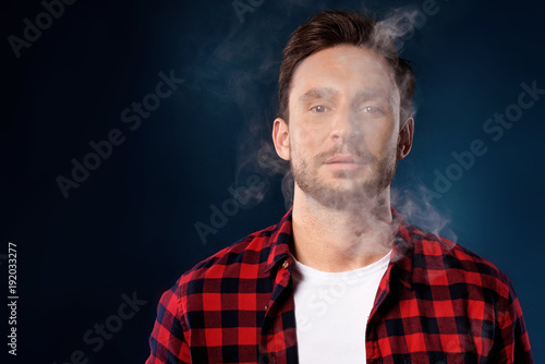 Handsome bearded man smoking electronic vape cigarette, Guy in a red t shirt smoking vapor, Man exhaled vapor on a dark studio background. Man smoking e-cigarette