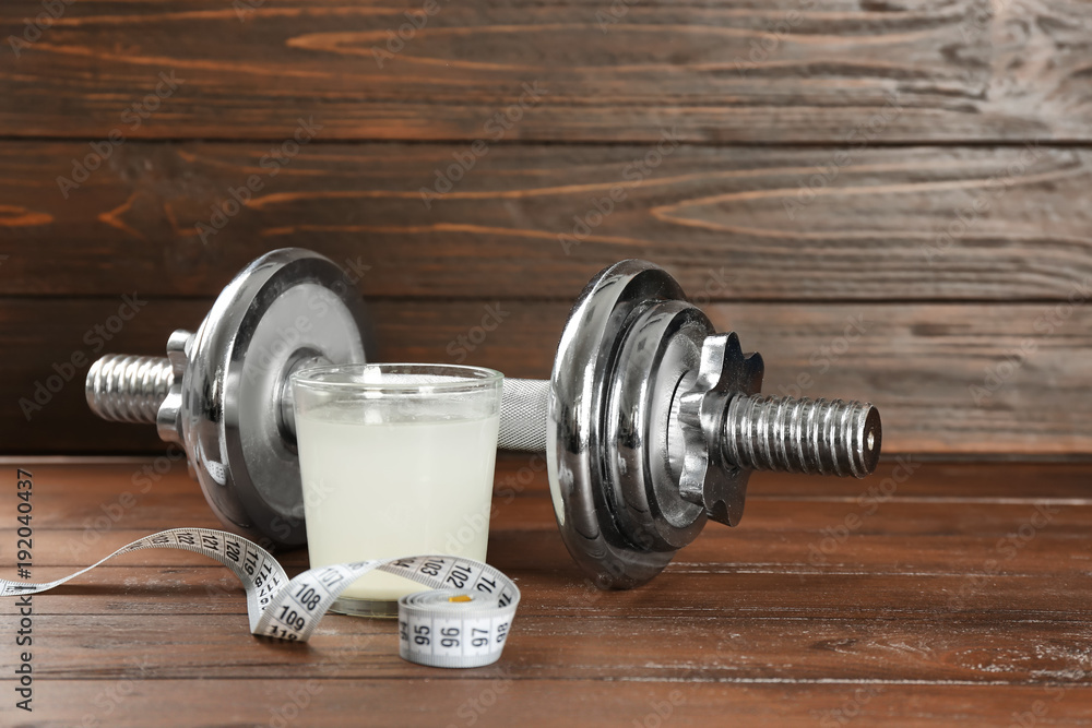 Glass with protein shake, measuring tape and dumbbell on wooden background Glass with protein shake, measuring tape and dumbbell on wooden background