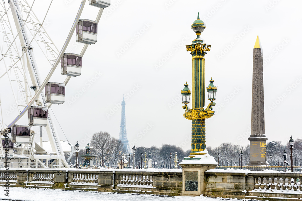 Winter in Paris in the snow. The Concorde square covered in snow with a decorated street light in the foreground, the Luxor obelisk and the Ferris wheel, and the Eiffel tower in the distance.