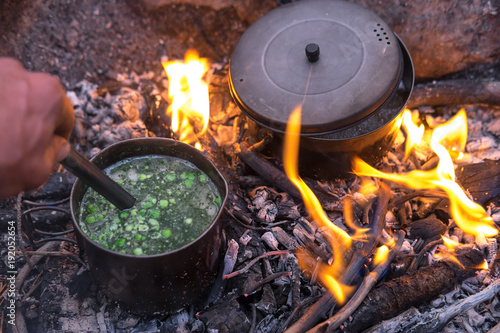 Two titanium pots cook food in a campfire. Flames from the fire move up the pans while a hand stirs soup with a spoon