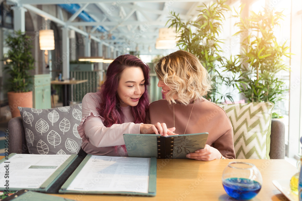 Two young women friends sitting in coffee shop or cafe and choose dishes. Female friendship, meeting togetherness Two young women friends sitting in coffee shop or cafe and choose dishes. Female friendship, meeting togetherness