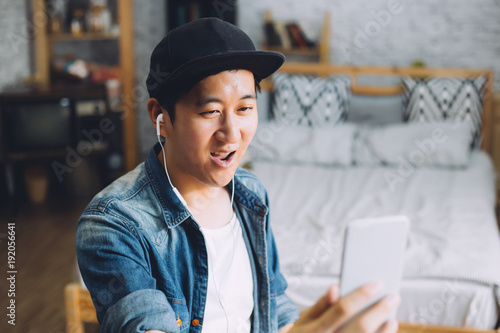 Young happy Asian man talking video call via smartphone wearing headphones at home