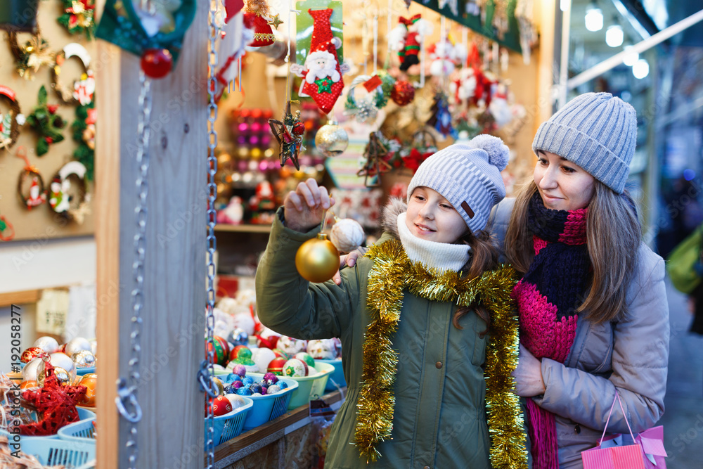 Smiling female and her daughter are choosing decorations for Christmas tree in the market outdoor.
