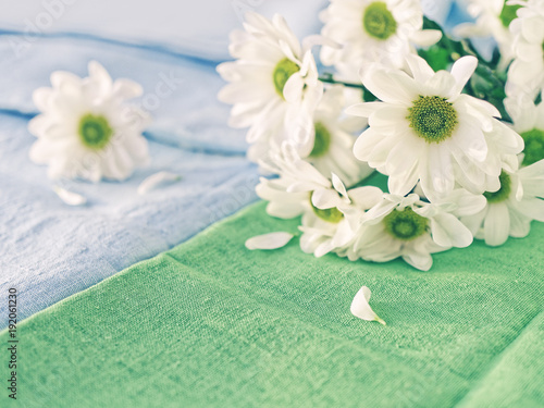 A bouquet of white chrysanthemum is on a green linen fabric. Nearby is a blue tablecloth with a flower on it. Cozy home background