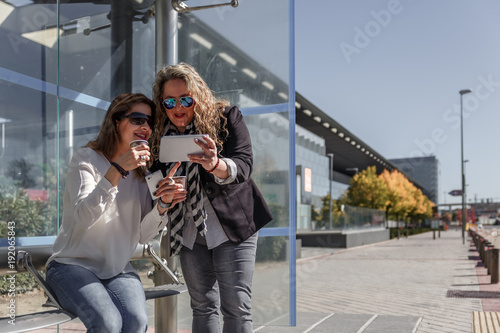 Two businesswomen with casual clothes carefully review a tablet, while they have a coffee and wait at a bus stop © Óscar
