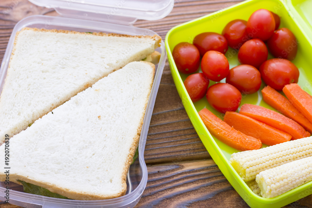 Fresh sandwich with lettuce in a plastic container and a green plastic container with baby corns, carrot and tomatoes, top view, dark board background Fresh sandwich with lettuce in a plastic container and a green plastic container with baby corns, carrot and tomatoes, top view, dark board background
