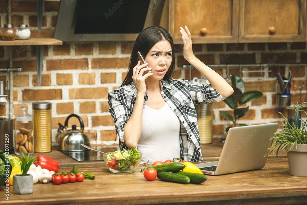 Laptop on kitchen table and cooking beautiful young woman. Food blogger concept. Using phone.