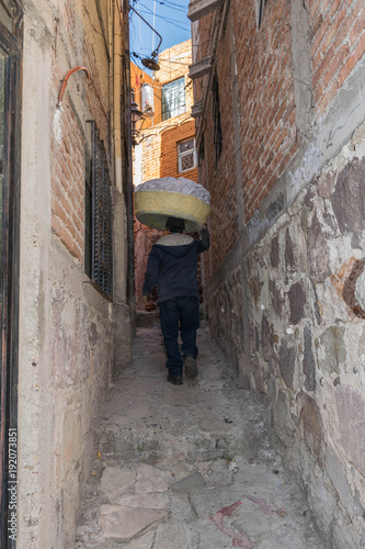 From the back, a man with a basket of laundry on his head, walking up a narrow stone pathway, between two stone walls, in Guanajuato, Mexico © Kathleen