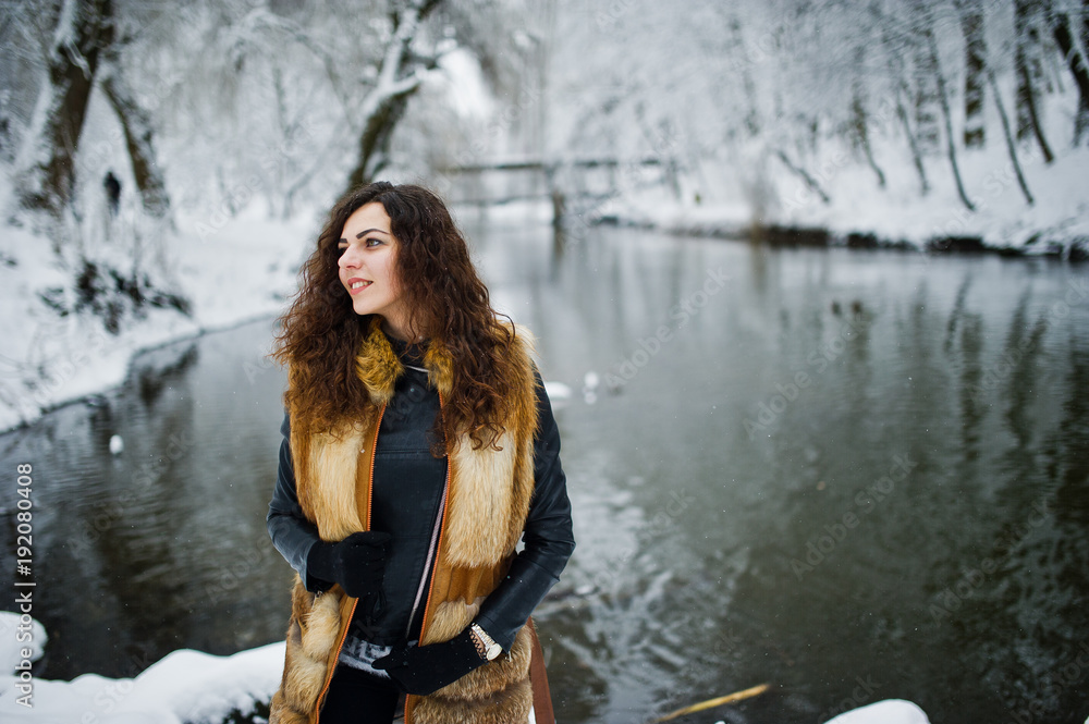 Elegance curly girl in fur coat at snowy forest park agasinst frozen river at winter.