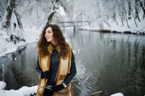 Fototapeta Naklejka Na Ścianę i Meble -  Elegance curly girl in fur coat at snowy forest park agasinst frozen river at winter.