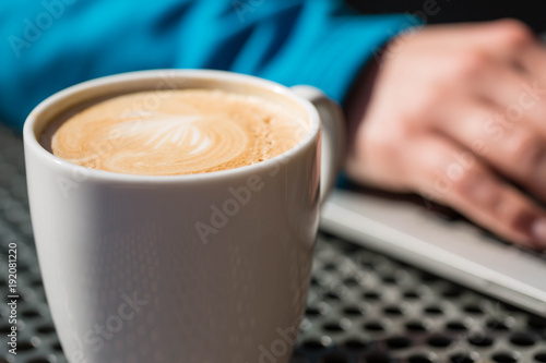 Coffee latte on a table from a cafe, with foam while a person works on a laptop computer