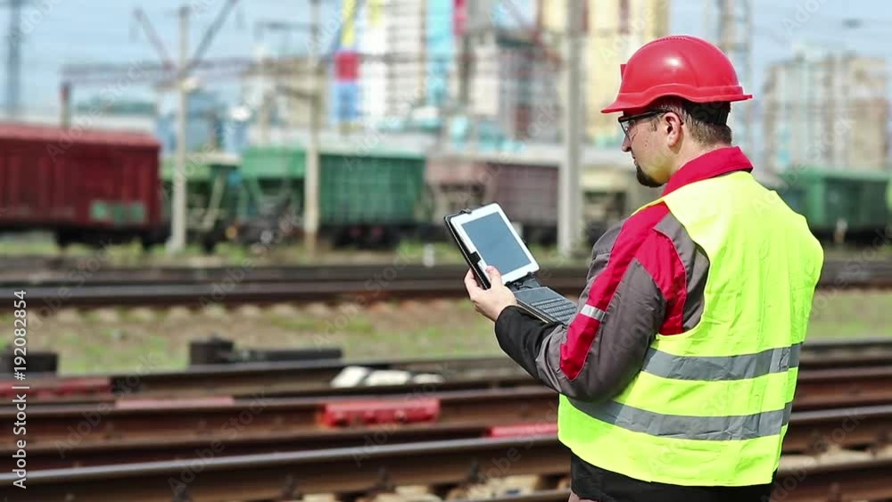 Railwayman with computer at freight train terminal.Railwayman in uniform and red hard hat works with tablet computer. Railway employee makes notes in his tablet pc Railwayman with computer at freight train terminal.Railwayman in uniform and red hard hat works with tablet computer. Railway employee makes notes in his tablet pc