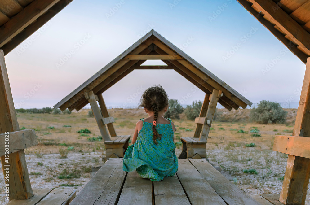 a small sad girl with a thin braid all alone in a green dress turned away and sits on a wooden table under the roof of an old altanka