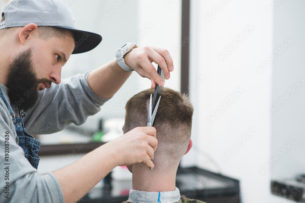 Fototapeta premium Portrait of a hairdresser with a beard who makes a hairdo to a young man. Creation of stylish men's hair scissors in the men's beauty salon. Haircut in barbershop