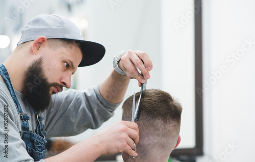 Portrait of bearded barber at work. Create a stylish men's hairstyle in barbershop. The hairdresser makes a hairstyle to the client with scissors and a comb