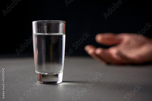 glass of vodka or alcohol drink, hand of a drunk man in the background, alcoholism and alcohol abuse concept, defocused, selective focus, close up, gray table, dark background