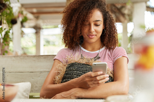 Image of African American female with curly hairstyle rests at cafe on couch huggs cushion, checks own email and messages with friends in social networks, uses mobile phone and wireless internet photo