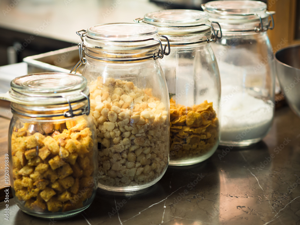 Glass bottles with cereals, crispy dice breads, chocolate chips, flour and icing sugar in a row on wooden table in kitchen or pantry for topping or ingredients of dessert or breakfast Glass bottles with cereals, crispy dice breads, chocolate chips, flour and icing sugar in a row on wooden table in kitchen or pantry for topping or ingredients of dessert or breakfast