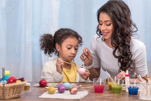 happy african american mother and daughter painting easter eggs