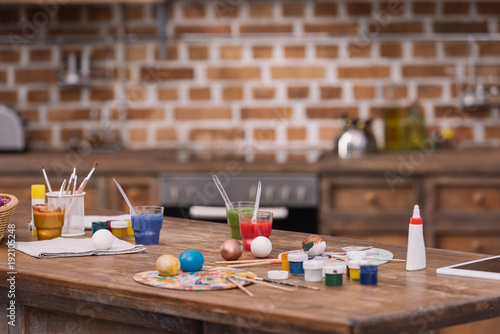 painted eggs with paints and brushes on wooden table in kitchen, easter concept