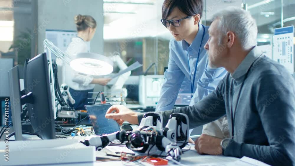 Two Engineers Talking while Working with Robot, Checking Data on Laptop Computer. In the Background Robotics Research Center Laboratory with Specialists Working.