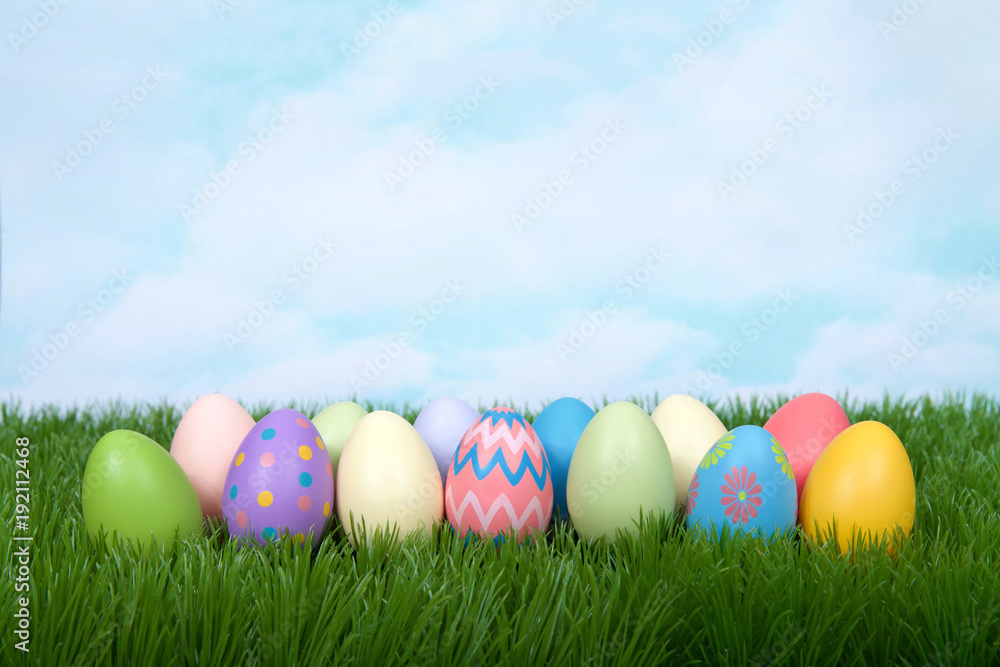 Colorful decorative and plain easter eggs lined up in a row in green grass. Blue sky with clouds in background.